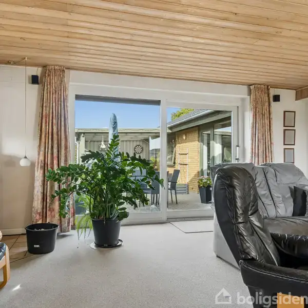 Brændeovn opvarmer rum med sorte læderstole og store grønne planter. Store glasdøre åbner ud til terrasse med siddemøbler og udsigt til træbeklædt loft og vægge.