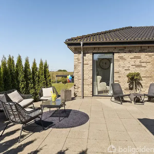 En terrasse med sorte havemøbler og et lille rundt bord står ved en murstensbygning. En hæk af høje buske omkranser området, og paraplyens skygge er synlig på fliserne.