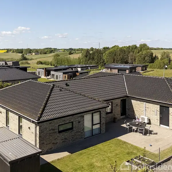 En moderne murstensvilla med sort tegltag står i en velplejet have. Et terrasseområde med havemøbler ses foran huset, omgivet af en grøn mark og flere huse i baggrunden.
