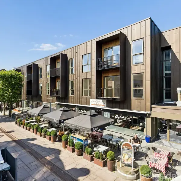 En moderne, brun treetages bygning med altaner står langs en gågade. Restauranter med udendørs servering, parasoller og planter fylder fortovet. Sollys kaster skygger på facaden.