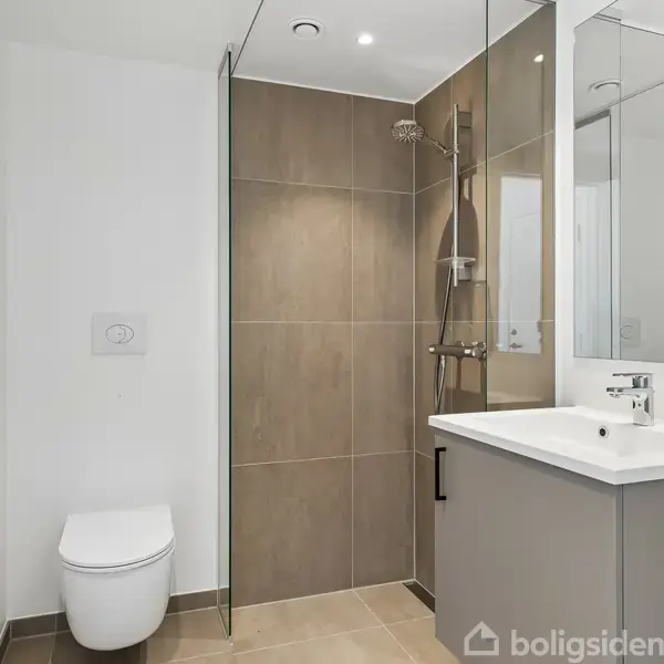 Et moderne badeværelse indeholder en væghængt toilet, en brusekabine med glasvægge og en stor flisevæg. Ved siden af bruseren er en håndvask med spejl over. Alt er i neutrale farver.
