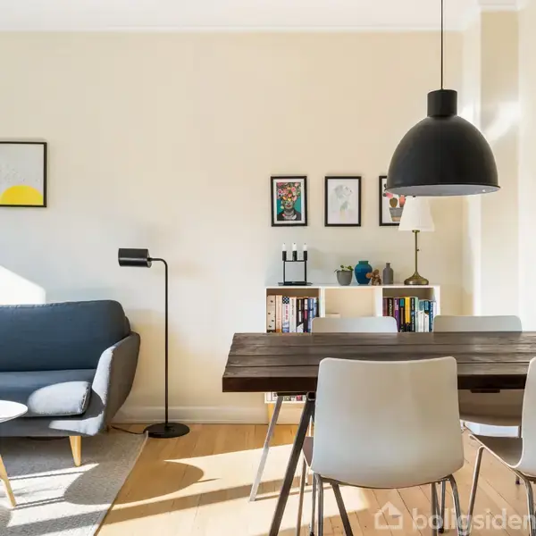 Sofa i hjørnet med en lampe ved siden af, ved siden af står et bord med hvide stole og en sort lampe over. Rummet er lyst med kunst på væggene.