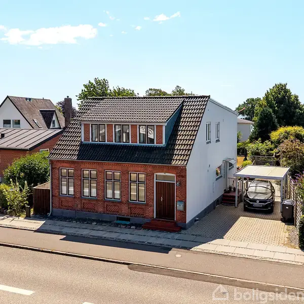 Et to-etagers hus med røde mursten og sort tag står ved en vej. Til højre er en indkørsel med en carport og en bil. Omkring ligger flere huse.