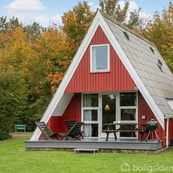 Rødt A-formet hus står stille, omgivet af efterårsfarvede træer. Foran huset er der en terrasse med stole og en grill. Græsplæne strækker sig foran.