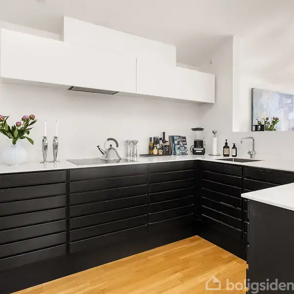 Sort-hvidt køkken med moderne design; sorte skuffer og hvide skabe. På bordet står blomster og flasker. Rum med trægulv og stort vindue, der giver naturligt lys.