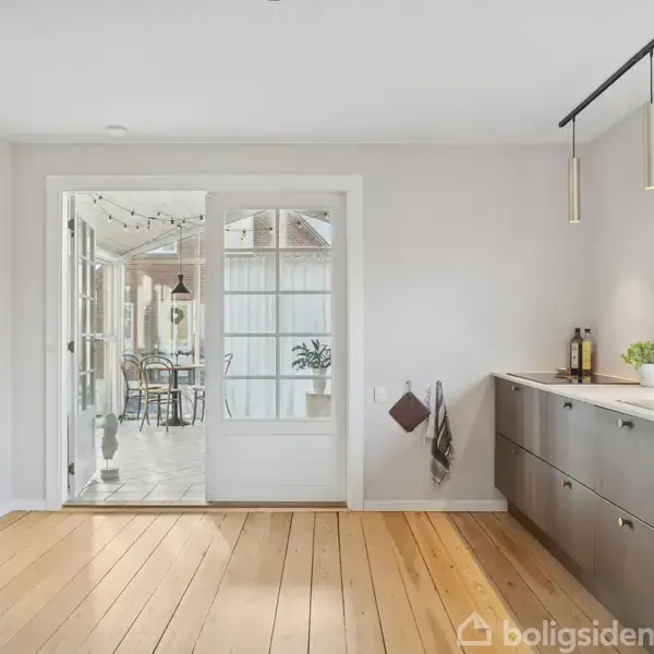 Køkken med lyse træplanker på gulvet, moderne skabe på højre side, og glasdøre der fører til en udestue. Udestuen har en glasvæg med udsigt til haven.