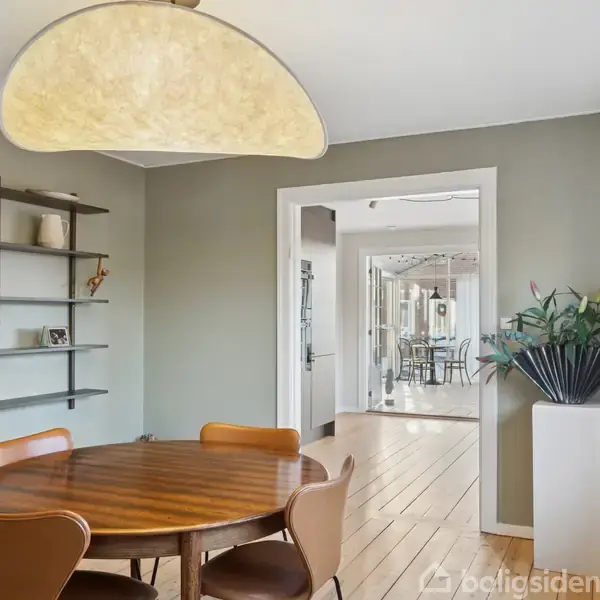 Et rundt træbord omgivet af stole står i en spisestue med grønne vægge. En stor lampe hænger over bordet. Til venstre er en væghylde, og en åbning fører til næste rum.