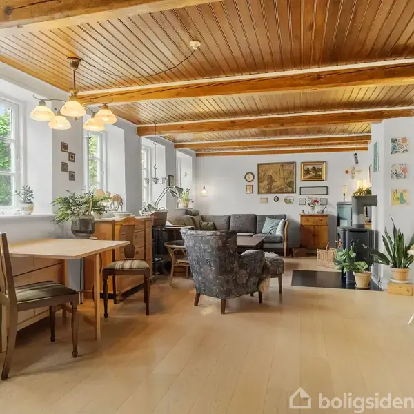 Stue med trægulv og træloftsbjælker, der indeholder spisebord med stole til venstre, sofahjørne med grå sofa og lænestol, planter og kunst på væggene, lys belysning, hygge og varme.