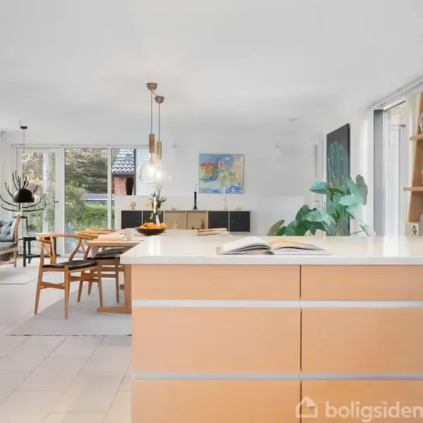 Et lyst køkken med træskabe har en åben bog på bordpladen. I baggrunden ses en spiseplads med stole, store vinduer og planter, der skaber en lys atmosfære.