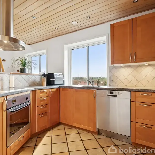 Et moderne køkken med træskabe og hvide fliser som backsplash. Rummet indeholder en ovn, opvaskemaskine og kaffemaskine. Sollys strømmer ind gennem store vinduer, hvilket skaber en lys og indbydende atmosfære.