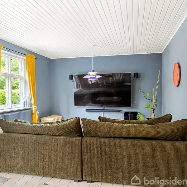 En stue med en grøn sofa vender mod et stort vægmonteret TV. Rummet har blå vægge, gule gardiner, en plante i hjørnet og et rundt, orange ur.