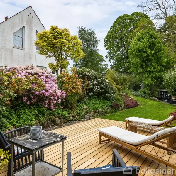 Træterrasse med to liggestole og bord, omgivet af frodig have med blomstrende buske og høje træer. Hvidt hus i baggrunden under en blå himmel.