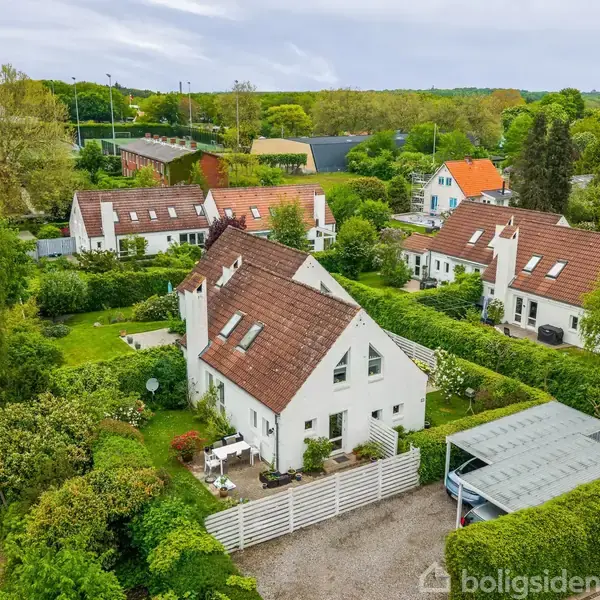 Hvide huse med røde tegltage ligger tæt sammen i en frodig, grøn boligforening. Små haver omgiver husene, og der er en parkeringsplads med et halvtag. Træer og buske fylder landskabet.