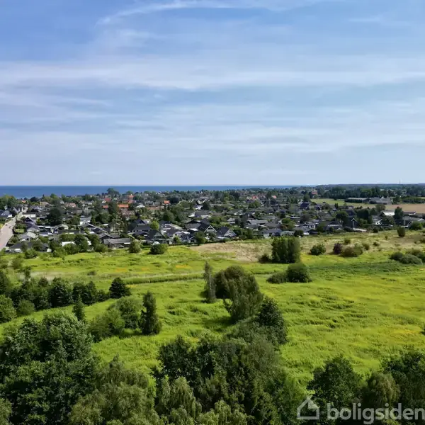 Et åbent grønt landskab strækker sig mod en kystby, hvor huse ses i horisonten. En vej skærer gennem området, mens havet anes i baggrunden.