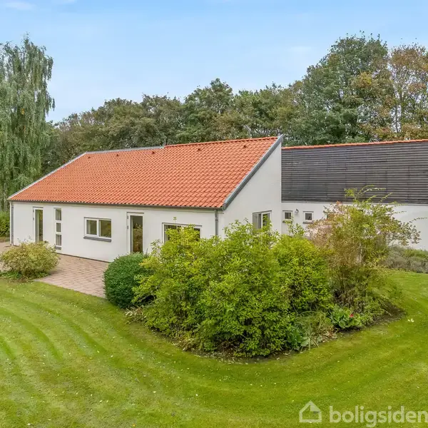 Et hvidt hus med rødt tag står på en frodig græsplæne omgivet af træer og buske, i en skovklædt have.