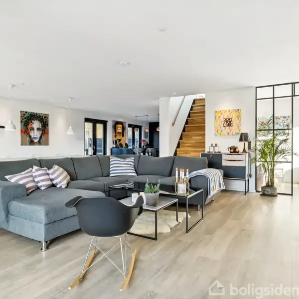 Stor grå sofa står i en moderne stue med lyse trægulve og kunst på væggene. I baggrunden ses en trappe og et glasadskilt rum.