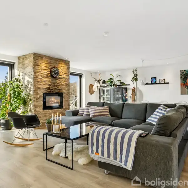 Gråt sofaarrangement med tæppe ligger i en stue med trægulv. Pejs på stenvæg, planter og kunst dekorerer rummet. Store vinduer åbner ud mod grøn have.