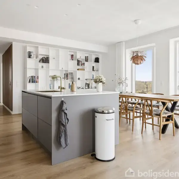 Køkken ø med gråt skab og vask, omgivet af moderne, lyse indretning med spisebord og stole ved store vinduer med udsigt. Hylder med bøger og dekorationer ses i baggrunden.