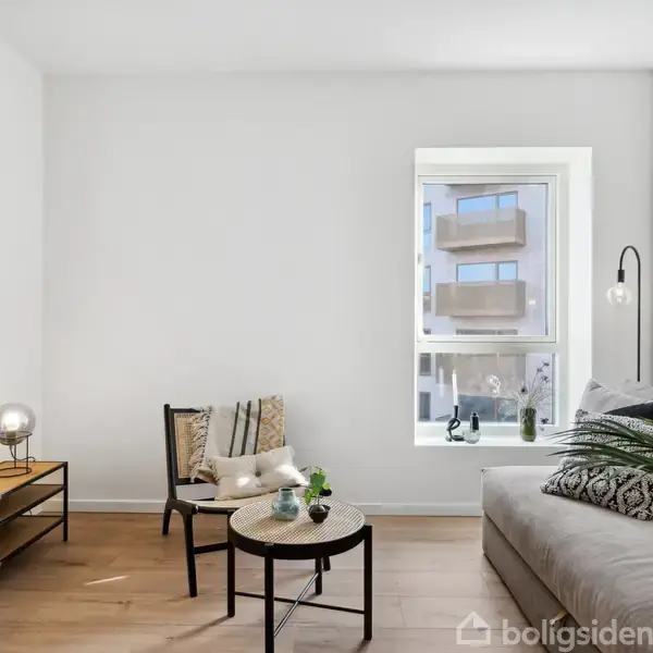 En moderne stue med en grå sofa ved siden af et stort vindue. Der er et lille rundt bord med planter foran sofaen, og en reol med en lampe og kunstplakat ved siden af.