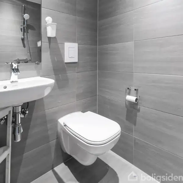 Et moderne badeværelse med en væghængt toiletkumme, håndvask med spejl og grå fliser på væggene. En flade hylde er installeret under vasken, og der er toiletpapirrulleholder på væggen.