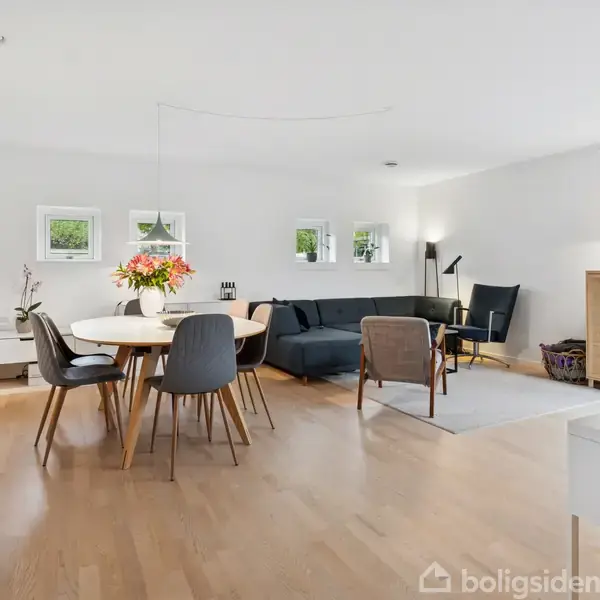 Spisestue med rundt træbord omgivet af stole, står i et lyst rum med trægulv. En grå sofa og lamper i baggrunden, skabe og reoler langs væggene.