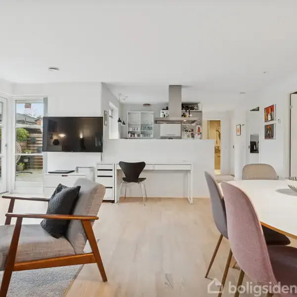 En lys stue med moderne møbler; en lænestol og et spisebord med stole. Et TV hænger på væggen. Store vinduer giver udsigt til en terrasse. Ingen tekst fundet.