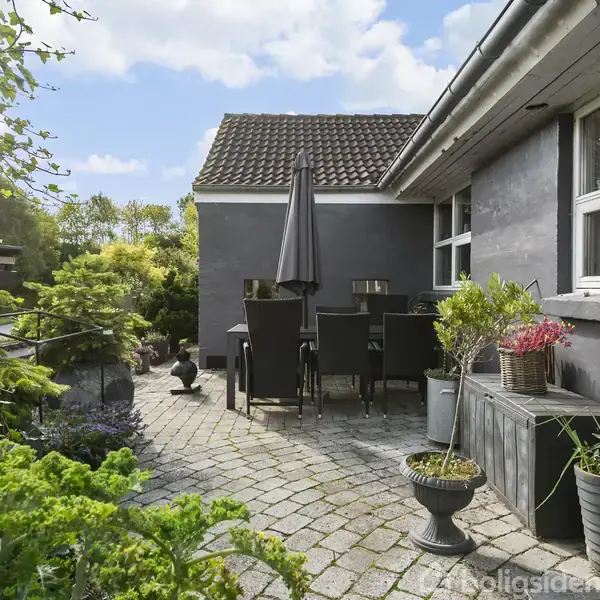 Terrassemøbler med parasol står på en flisebelagt terrasse. Omkring er der grønne planter og blomster i krukker ved siden af et gråt hus. Himlen er blå med hvide skyer.