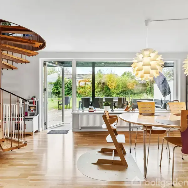 En moderne spisestue med et rundt træbord omgivet af stole. En spiraltrappe til venstre fører til en øvre etage. Store vinduer åbner op til en have udenfor.