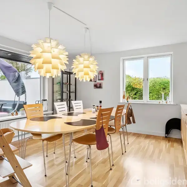 Et moderne spisestuebord med seks træstole står under to dekorative pendellamper. Rummet er lyst og rummeligt med stort vindue og udsigt til en grøn have.