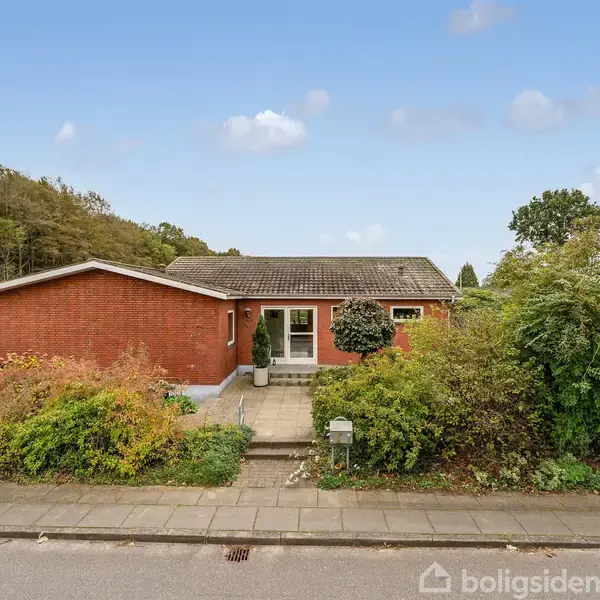 Et parcelhus i rød mursten står ved vejen, omkranset af en frodig have. Baggrunden domineres af træer under en klar blå himmel.