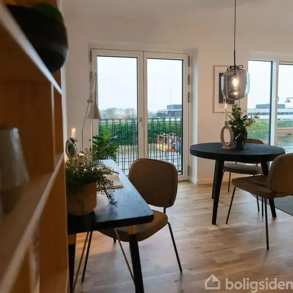 Stue med træmøbler står i et lyst rum. Rundt spisebord med fire stole står nær vinduer, som åbner ud til en balkon. Planter dekorerer rummet.