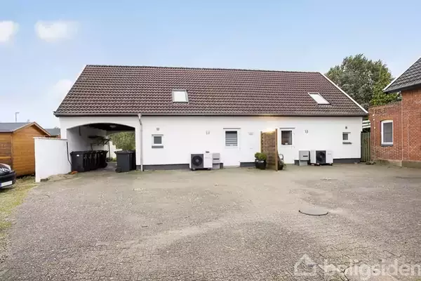 Et hvidt hus med brunrødt tag, står på en stor gårdsplads. Der er to trapper op til indgange og to tagvinduer. Til venstre er en carport.