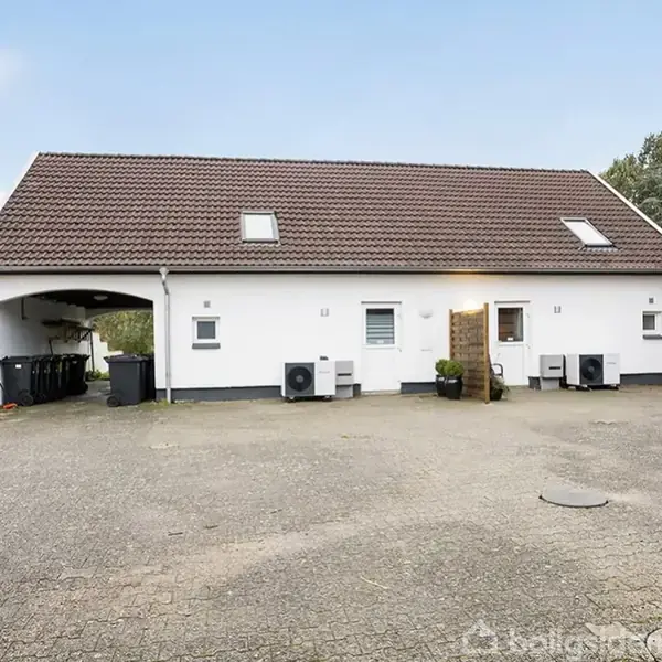 Hvidt hus med mørkt tag står stille. Gårdsplads foran med flisebelægning og indkørsel. Til venstre ses en carport med affaldscontainere. Bag huset er der træer.