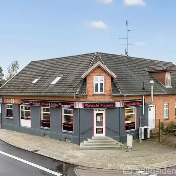 En rød murstensbygning med skifertag huser "Kerimh Pizzeria". Bygningen har flere vinduer og en hovedindgang med trappe. Omkring er der fortov og en vej.