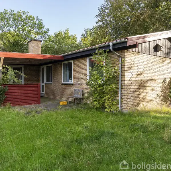 Et enkelt etplanshus af mursten med rødt tag og skorstensrør, beliggende i en frodig have med græs og træer omkring. En lille veranda med rødt hegn ses foran.