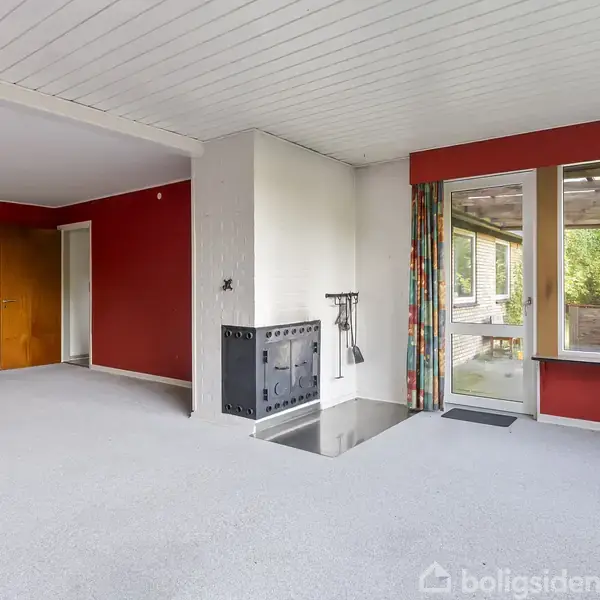 Stue med åben pejs i et hjørne, pejsesæt hænger ved siden af. Store vinduer med farverige gardiner. Røde vægge, hvidt loft, åbner ud mod en grøn have.