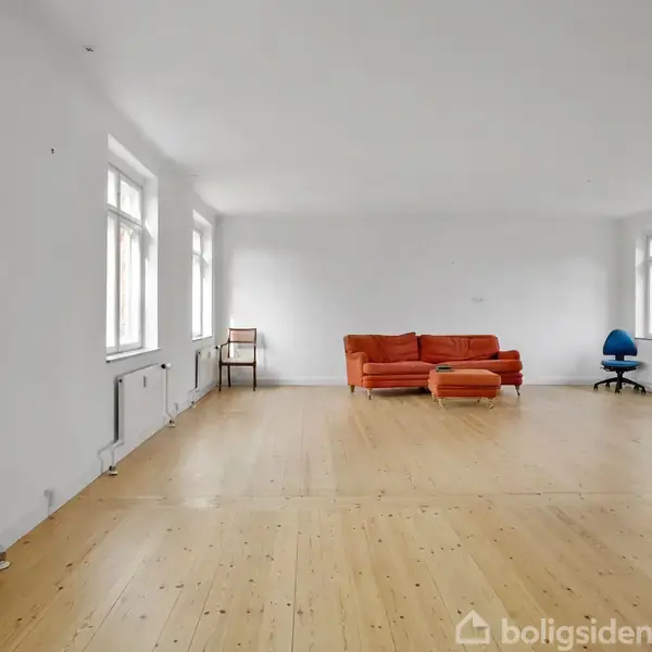 En orange sofa står i et rummeligt, lyst rum med trægulv og hvide vægge. I nærheden ses en blå kontorstol og et lille bord.