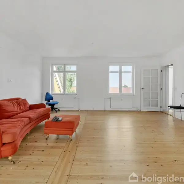 Rød sofa står i en lys, rummelig stue med trægulv. En blå kontorstol er placeret ved et vindue. Der er to vinduer og en dør i baggrunden.