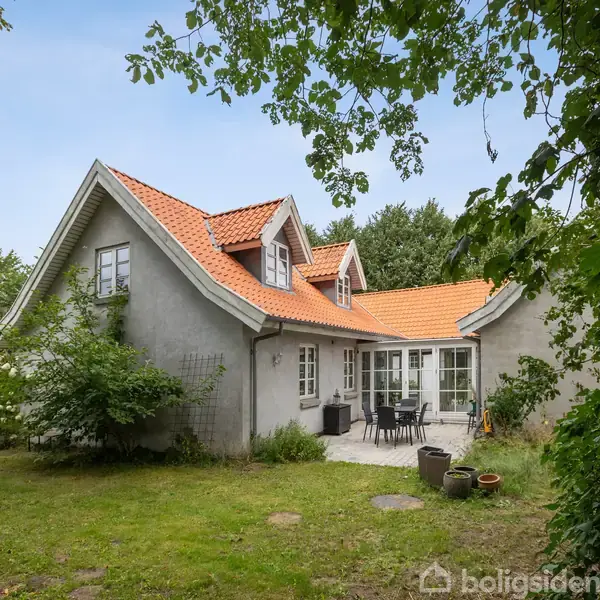 Huset har hvide vægge og rødt tegltag, med kviste. Der er en terrasse med havemøbler. Omgivet af grønne træer og græsplæne i en landlig have.