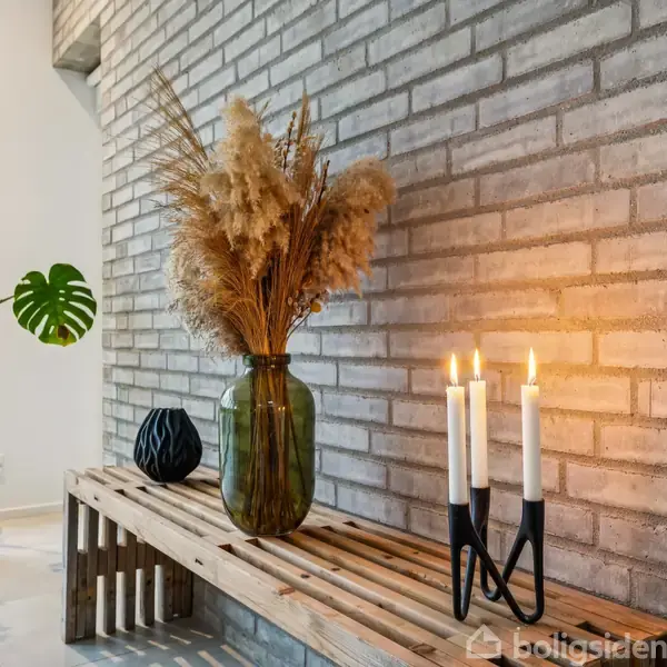 En stor grøn vase med tørrede siv står på en træbænk ved en murstensvæg. Ved siden af er tre tændte stearinlys i en holder. En plantepotte er i baggrunden.