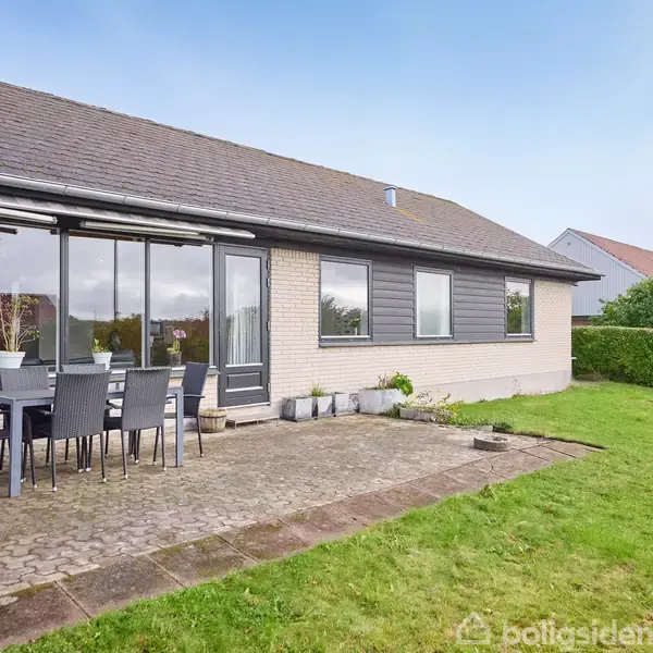Hus i én etage med hvid murstensfacade og grå detaljer. Terrasse med spisebord og stole i forhaven, omgivet af grøn græsplæne og hæk i en rolig villakvarter.