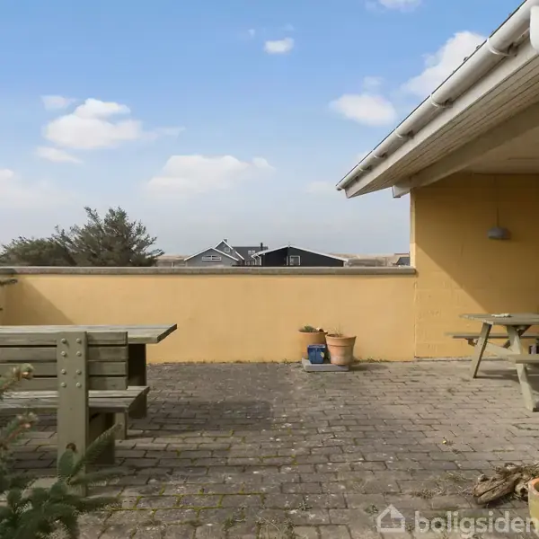 En flisebelagt terrasse med to træbænke og bord, omgivet af gule vægge, og udsigt over nabolaget med huse og himmel i baggrunden. Der står også nogle krukker og en grill.