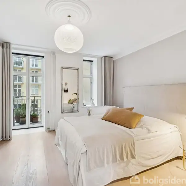 En dobbeltseng med hvidt sengetøj og pudebetræk står i et lyst soveværelse med trægulv. To store vinduer med lange gardiner giver udsigt til en bygning udenfor.