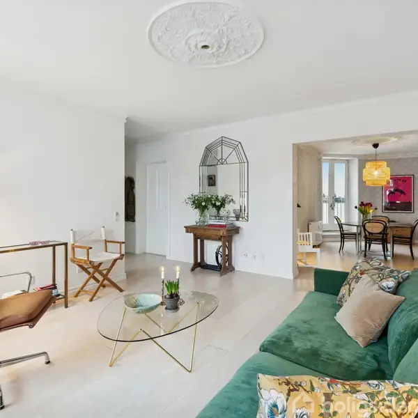 En grøn sofa står i en lyst indrettet stue med et rundt glasbord foran. Stuen har moderne møbler, en brun lænestol, og en åbning til spisestuen med flere stole og et spisebord.