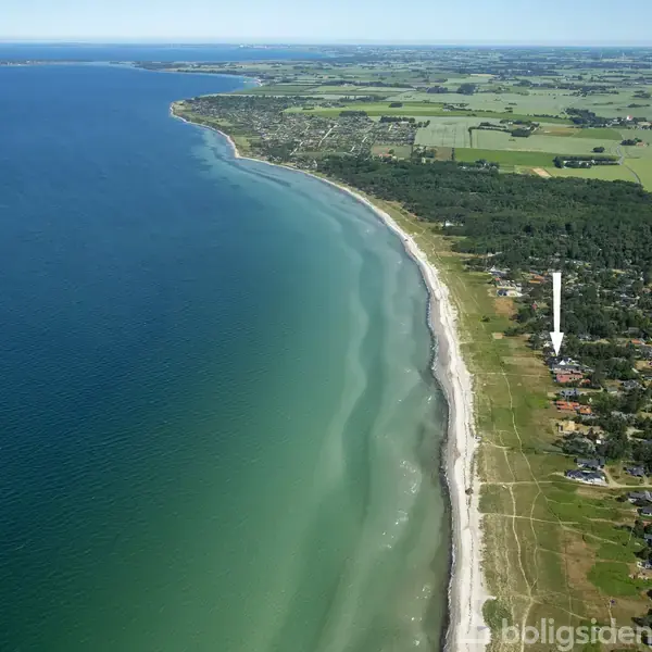 Kystlinje strækker sig langs en bred sandstrand med klart blåt vand. I baggrunden ligger grønne marker og en række huse. En hvid pil peger mod et bestemt husområde.