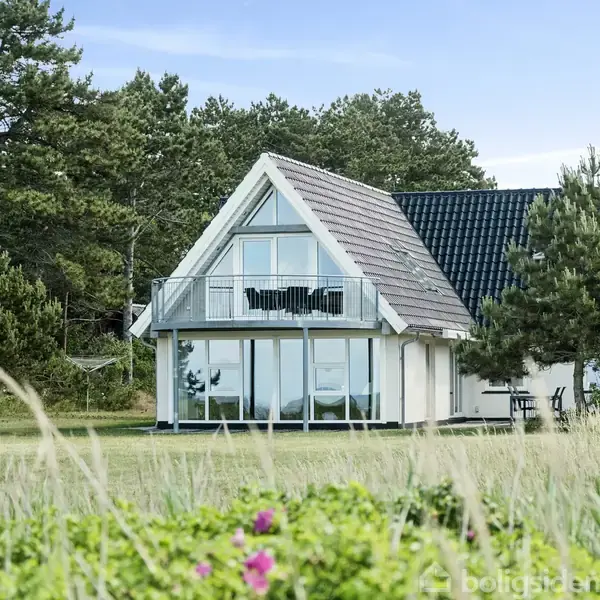 Hus med trekantet gavl og balkon ses fredeligt stående omgivet af træer og buske i en grøn have, under en blå himmel.