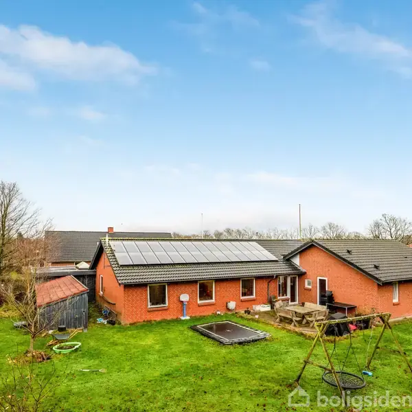 Rødt murstenshus med solpaneler på taget, står i en grøn have med gyngestativ og trampolin. Huset har hvidmalede vinduer og døre, beliggende i et villakvarter.