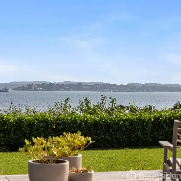 Terrassemøbler står stille foran smukke blomsterpotter. I baggrunden ses en blå fjord og en skovbeklædt horisont, skaber en fredelig og naturskøn atmosfære.