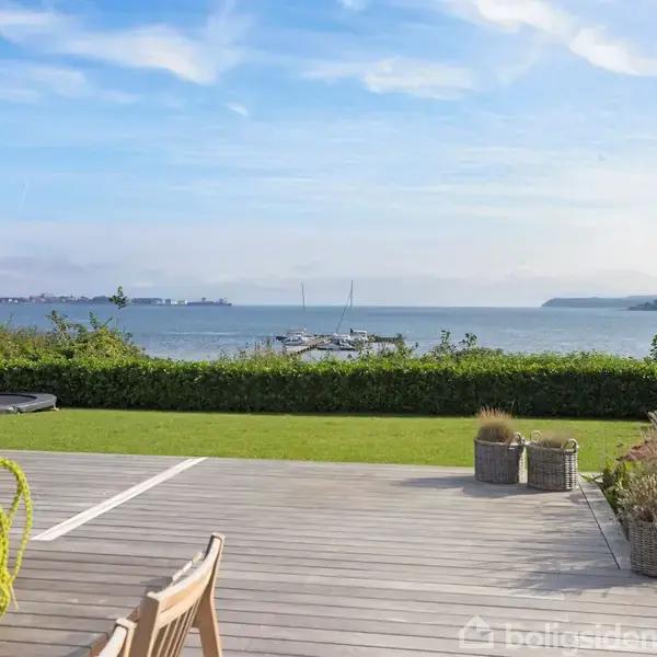 Terrassen er udstyret med træmøbler, omkranset af dekorative planter, og præger en udsigt over en grøn have og havet i baggrunden. Himmel med lette skyer ses også.