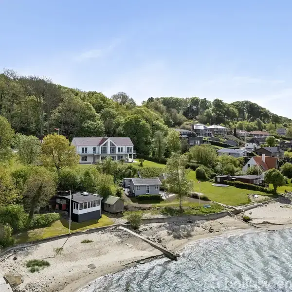 Stort hvidt hus ligger på en bakketop omgivet af træer. Handling: Det står stille. Kontekst: Strandsamfund med vand og huse, grønne områder og en lille kystlinje.